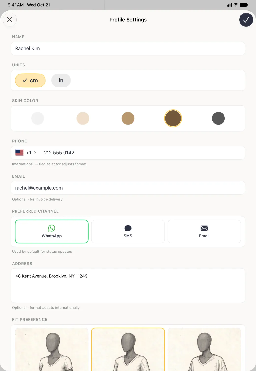 Profile Settings sheet covering the screen below the status bar, with a cross at the top left and a navy checkmark at the top right, the name Rachel Kim in a white card under NAME, cm and in buttons under UNITS with cm ticked, five skin tone circles under SKIN COLOR with brown ringed in gold, then PHONE +1 212 555 0142, her email under EMAIL, PREFERRED CHANNEL with WhatsApp outlined in green, the ADDRESS 48 Kent Avenue, Brooklyn, NY 11249 and the top of the FIT PREFERENCE cards with Regular outlined in gold.